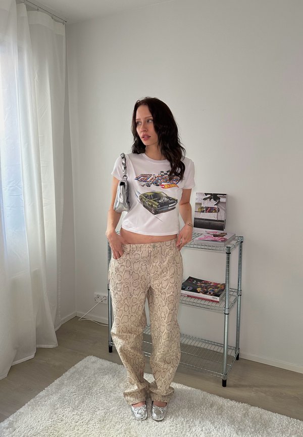 Λευκό t-shirt με γραφιστικό σχέδιο αυτοκινήτου, συνδυασμένο με μπεζ παντελόνι με σχέδιο από φίδι. Ασημένια παπούτσια slip-on ολοκληρώνουν το σύνολο. Μη μινιμαλιστικό φόντο.