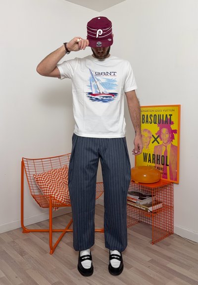 Hombre ajustándose una gorra granate, vistiendo camiseta blanca con gráfico, pantalones de rayas finas y zapatos en blanco y negro, de pie junto a una silla naranja y un póster colorido de Basquiat-Warhol.