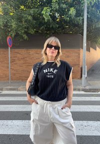 Melna svītraina bez piedurknēm augša ar baltajiem akcentiem, logo ar tekstu "NIKE 1972." Baltas kravas bikses, melna pleca soma un tumši saulesbrilles.