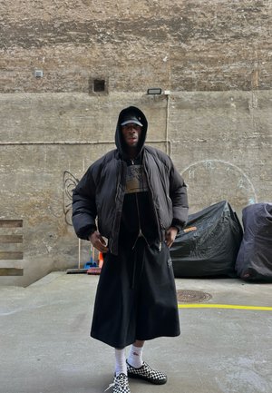 Hombre con chaqueta acolchada negra, capucha y gorra, de pie frente a una pared de hormigón texturizado con un reloj dibujado con tiza y objetos cubiertos cerca.