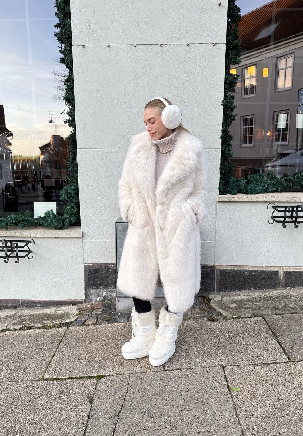 Lunga e folta giacca in pelliccia finta bianca sopra un maglione leggero, abbinata a cuffie bianche e stivali bianchi chunky con lacci. I leggings scuri completano l'outfit.