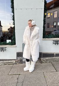 Lunga e folta giacca in pelliccia finta bianca sopra un maglione leggero, abbinata a cuffie bianche e stivali bianchi chunky con lacci. I leggings scuri completano l'outfit.