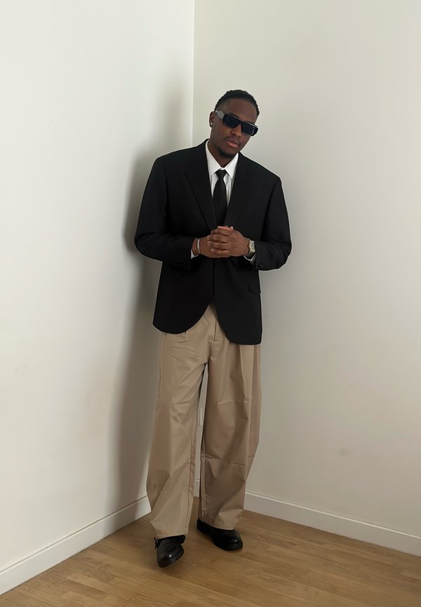 Homme portant un blazer noir, une chemise blanche, une cravate noire, un pantalon beige à jambes larges, des chaussures noires et des lunettes de soleil, debout devant un mur blanc uni.