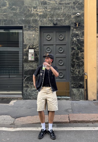 Camisa negra de manga corta con cremalleras, pantalones cortos de cargo beige, zapatos negros y calcetines blancos. Sosteniendo una bebida verde cerca de una pared de piedra texturizada y una puerta.