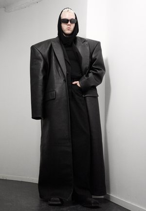 Persona con cabello corto rubio que lleva un abrigo de cuero negro de gran tamaño, sudadera con capucha negra, pantalones, zapatos y gafas de sol oscuras, de pie contra una pared blanca.