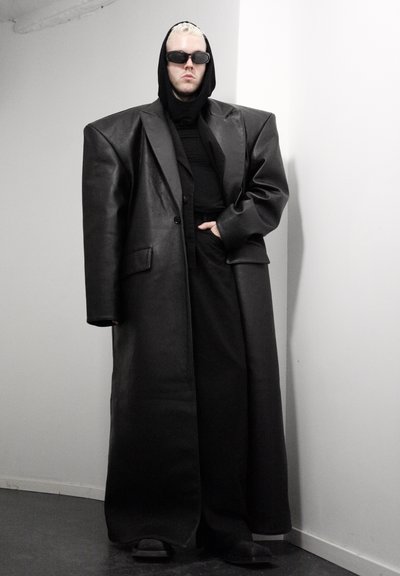 Persona con cabello corto rubio que lleva un abrigo de cuero negro de gran tamaño, sudadera con capucha negra, pantalones, zapatos y gafas de sol oscuras, de pie contra una pared blanca.