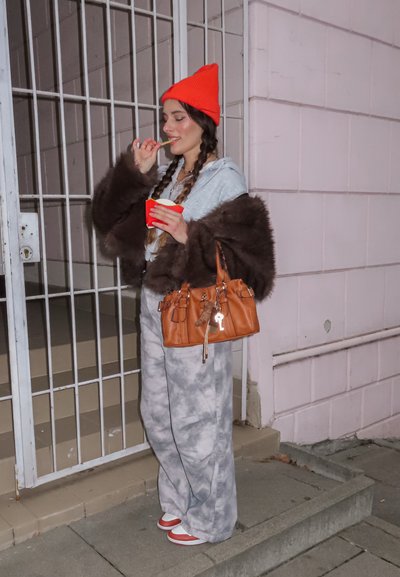 Persona con una sudadera con capucha gris y un abrigo de piel marrón, pantalones grises con estampado, gorro rojo y zapatos rojos, sosteniendo una taza roja frente a una puerta metálica.