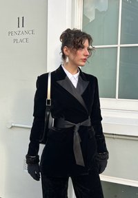 Blazer en velours noir avec revers en satin et ceinture assortie, porté avec une chemise blanche et des gants noirs. Fenêtre en arrière-plan.