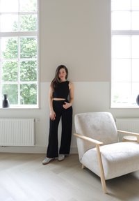 Fekete crop top és bő szárú nadrág, amelyet egy modell visel, aki egy világos, texturált karosszék mellett áll, fából készült akcentusokkal egy világos szobában.