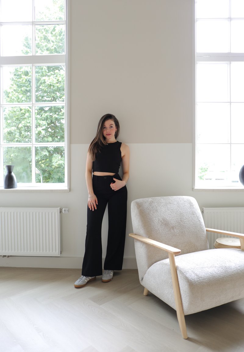 Μαύρο κροπ τοπ και παντελόνι με φαρδιά πόδια που φοράει ένα μοντέλο, στέκοντας δίπλα σε μια ανοιχτόχρωμη, υφασμάτινη πολυθρόνα με ξύλινα στοιχεία σε φωτεινό δωμάτιο.