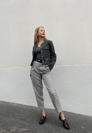 Chaqueta de tweed gris con botones dorados, superpuesta a una blusa gris, combinada con pantalones gris claro y mocasines oscuros sobre un fondo sencillo.