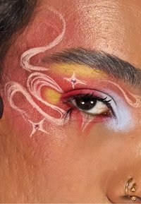 Närbild av ett öga med livfull makeup, med röda, gula och blå nyanser, virvlande mönster och glittriga detaljer, samt en guldig näsring.