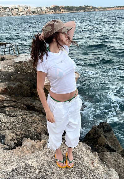 Camiseta blanca corta con gráfico, pantalones blancos sueltos, chanclas amarillas y verdes, y una gorra de camuflaje. El fondo presenta una costa rocosa y agua.