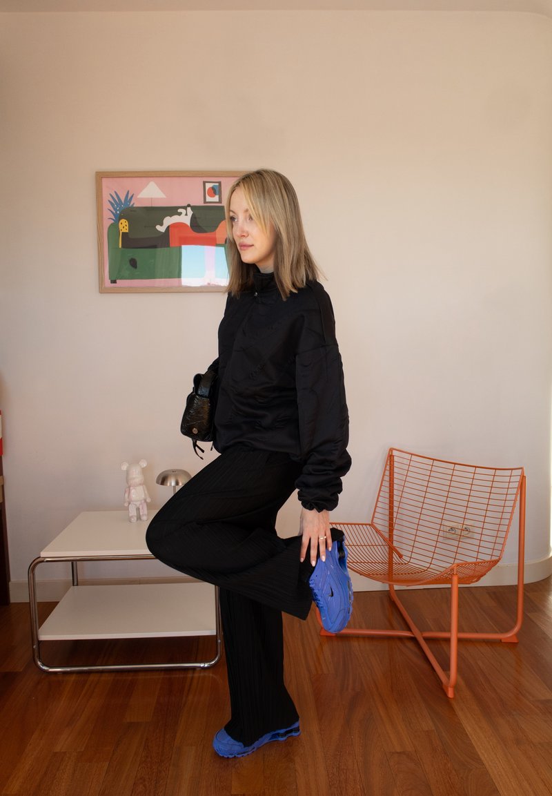 Femme en tenue noire ajustant sa chaussure bleu vif à l'intérieur, avec une chaise en fil orange moderne, une table blanche et des œuvres d'art murales colorées derrière elle.