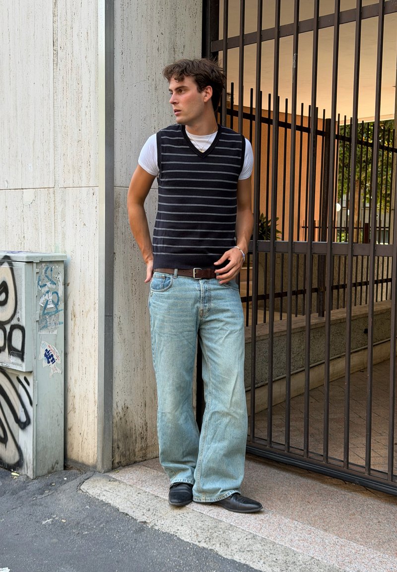 Ung mand iført en stribet sort vest over en hvid t-shirt, lyseblå jeans og sorte sko, står ved en metalport og en væg med graffiti.