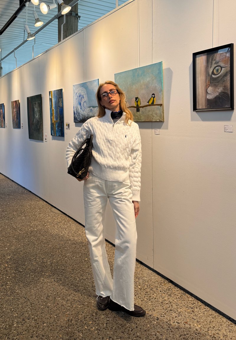 Frau in weißem Pullover und Hose hält eine schwarze Tasche und steht in einer Kunstgalerie mit Gemälden von Wellen, Vögeln und einer Katze an der Wand.