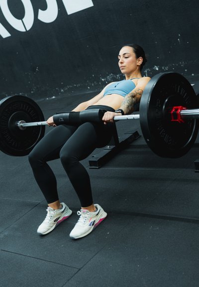 Barra de pesas con pesas sostenida por una persona que lleva un sujetador deportivo de color azul claro y leggings negros, sobre un suelo de gimnasio negro con una pared gris detrás.