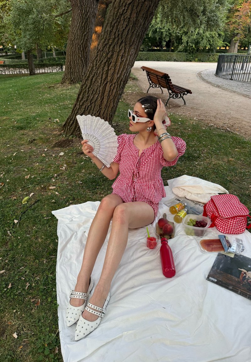 Haut à carreaux rouges avec des manches volantes, éventail blanc, chaussures blanches à clous, pique-nique sur une couverture blanche avec des fruits et des boissons sur l'herbe.