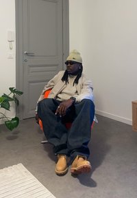 Oranssi tuoli, harmaa seinä ja ruskea lattia. Henkilö, jolla on beige pipo, aurinkolasit, harmaa takki ja tummansiniset farkut sekä vaaleat saappaat. Sisäkasveja näkyvissä.