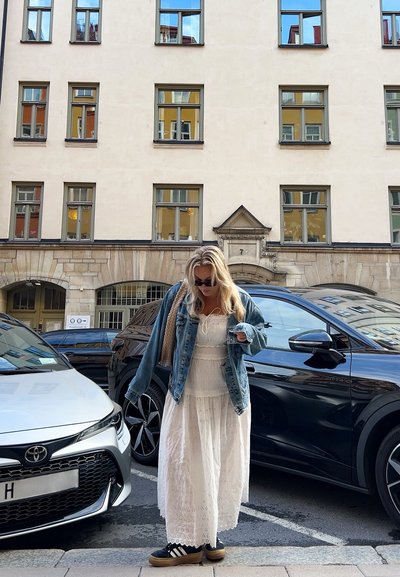 Chaqueta de mezclilla sobre un vestido maxi blanco bordado, zapatillas negras con suelas de plataforma, de pie entre dos coches estacionados en una calle de la ciudad.