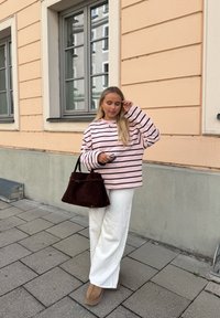 Stribet lyserød sweatshirt, hvide bukser med vide ben, brune ruskindssko og en mørkebrun shoppertaske. Står op ad en lysefarvet væg.