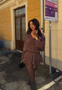 Blouse marron fluide avec ourlet à volants, associée à des collants transparents marron à imprimé léopard et des bottines noires. Sac à main noir et bijoux argentés en accents.