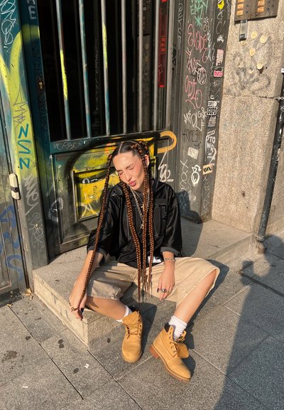 Camisa negra de gran tamaño con letras en negrita, pantalones cortos de carga beige y botas de cordones marrones. Se pueden ver trenzas largas y anillos de plata.