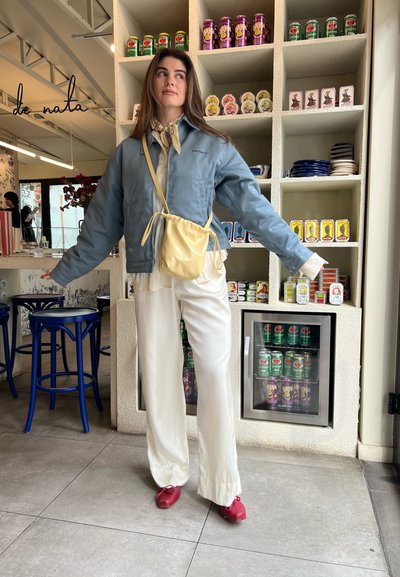 Chaqueta bomber azul, blusa blanca, pantalones de pierna ancha crema, bolsa amarilla con cordón, zapatos rojos; entorno de cafetería interior con latas de bebidas en el fondo.