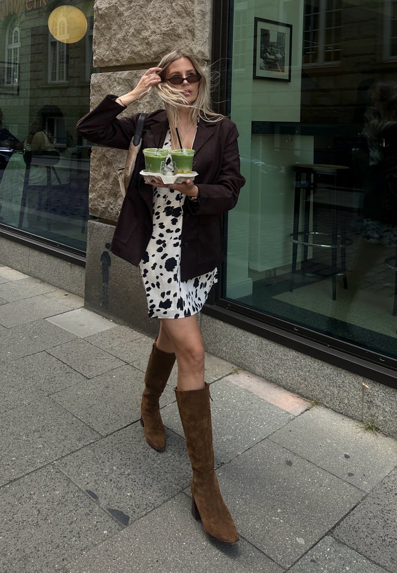 Modèle tenant deux boissons vertes dans un plateau en papier, portant un blazer noir par-dessus une robe blanche à pois noirs et des bottes en daim marron jusqu'aux genoux.