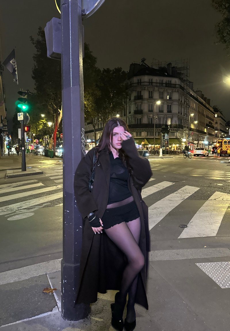 Vrouw in een zwarte jas, shorts en tights leunend tegen een straatpaal op een stads kruisingspunt bij nacht met verlichte gebouwen.