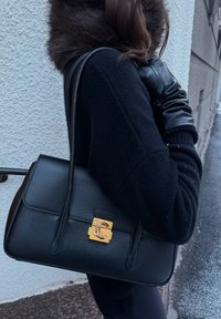 Personne portant un manteau noir et des gants en cuir tient un sac à main en cuir noir avec un fermoir doré, debout près d'un mur blanc texturé en plein air.