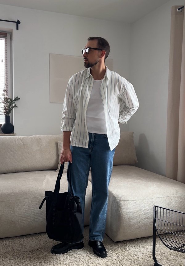 Hombre con barba y gafas, vestido con camisa de rayas y vaqueros, de pie en la sala de estar sosteniendo una bolsa negra junto a un sofá beige.
