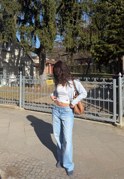 Mujer con vaqueros azul claro, top corto blanco y cárdigan, de pie en la acera cerca de una valla metálica con árboles y casas bajo la luz brillante del día.