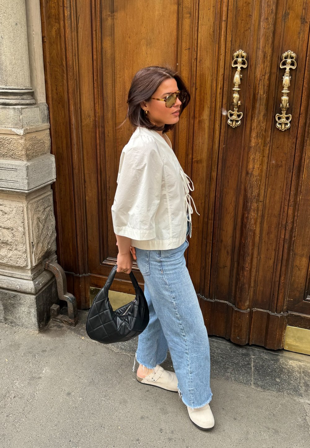 Λευκή κοντή ζακέτα με κορδόνια, ανοιχτό μπλε φαρδιές παντελόνες με ακανόνιστο τελείωμα, λευκά slip-on παπούτσια και μια μαύρη τσάντα με κουβερτίνα.