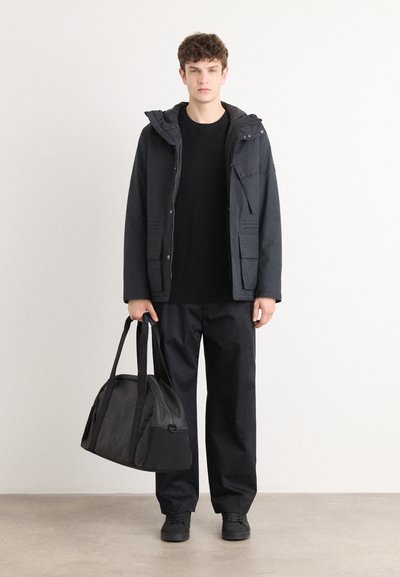 Chaqueta de plumas negra con capucha, llevada sobre un suéter negro texturizado y pantalones negros holgados. Lleva un bolso de deporte negro. Zapatos negros.