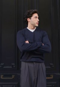 Granatowy sweter w serek z długimi rękawami, zestawiony z szarymi spodami plisowanymi, na tle czarnej panelowej ściany.