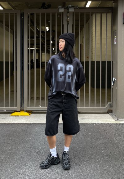 Jersey negro oversize con el número 22, acompañado de pantalones cortos de mezclilla negros. Zapatillas negras y calcetines blancos completan el look. Puerta gris en el fondo.
