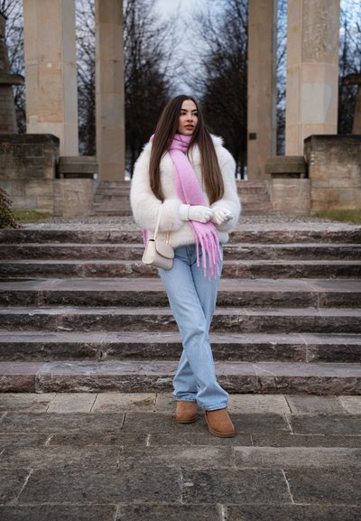 Suéter blanco esponjoso, bufanda rosa con flecos, vaqueros azules y botas de tobillo marrones. Un bolso gris y guantes completan el conjunto. Escaleras de piedra de fondo.