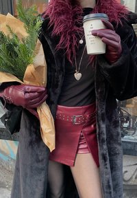 Black faux fur coat, burgundy leather mini skirt adorned with studs, matching gloves, holding an evergreen wrapped in paper, and a coffee cup with printed text.