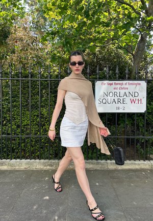 Mujer con gafas de sol y conjunto beige caminando junto a un cartel de la calle Norland Square con bolso negro y sandalias negras en un día soleado.