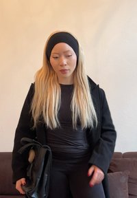 Manteau noir, haut noir ajusté et pantalon noir. Longs cheveux blonds et un bandeau noir. Tient un sac à main noir avec un détail en pompon en fourrure.