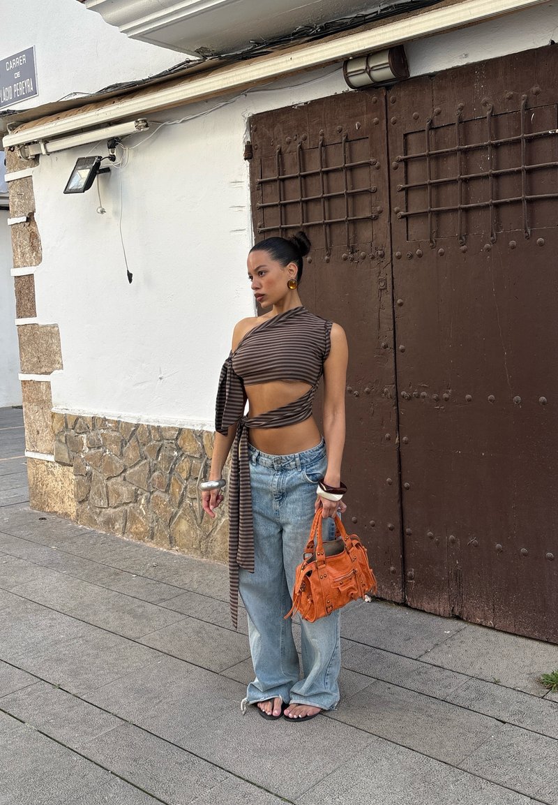 Femme avec un crop top rayé à une épaule et un jean ample, tenant un sac à main orange, debout contre un mur en pierre et blanc avec de grandes portes marron.