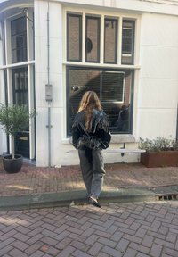 Fekete, fényes, redőzött dzseki, megkötve hátul, szürke nadrág felett. Téglával burkolt utcán áll egy nagy ablak és egy cserepes növény közelében.
