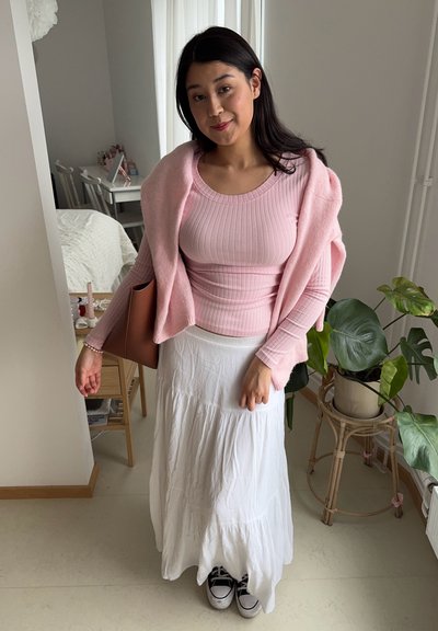 Suéter de canalé rosa claro y blusa de manga larga a juego combinados con una falda blanca fluida. Bolso marrón, zapatillas negras, vegetación de fondo.