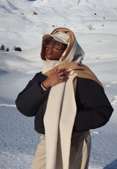 Persona con gafas de sol, gorra beige, bufanda bicolor beige y crema que cubre la cabeza, chaqueta negra y anillos, de pie en un terreno montañoso nevado.