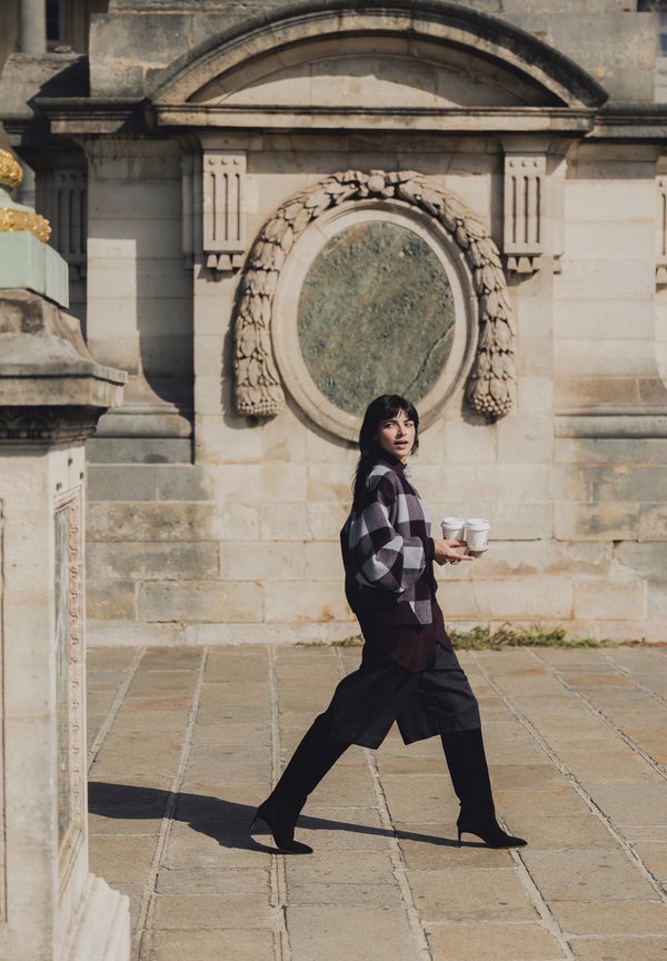 Femme en pull à carreaux et bottes noires marchant en plein air, tenant deux tasses de café dans une place historique en pierre ornée de décors muraux.