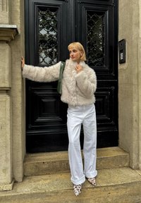 Femme aux cheveux blonds courts portant une veste en fausse fourrure beige, un pantalon blanc et des chaussures pointues à motifs, debout près d'une porte noire avec des panneaux de verre ornés.