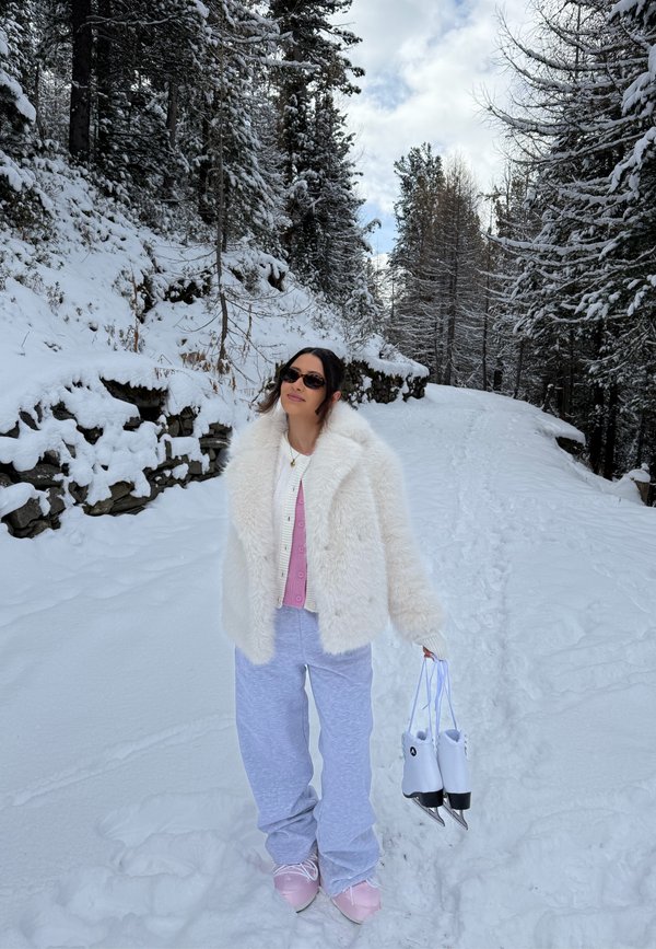 Chaqueta de piel sintética blanca sobre un suéter rosa y pantalones anchos de color gris claro. Sosteniendo patines de hielo blancos en un entorno boscoso nevado.