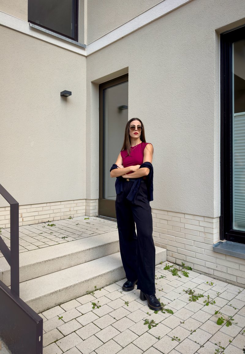 Uma mulher com uma blusa ajustada bordeaux e calças largas pretas está em degraus. Ela usa óculos de sol e tem um casaco azul-marinho pendurado sobre os braços.