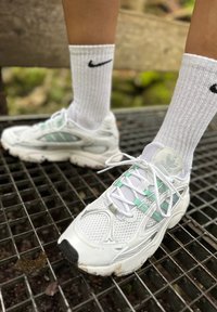 Bijele atletske tenisice s mrežastim detaljima, zelenim akcentima i bijelim vezicama. Nosi se s bijelim rebrastim Nike čarapama. Postavljene na rešetkastu površinu.